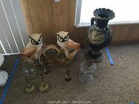 Photo showing two ceramic owl figures with a central clock, two small brass candle holders, and glass vase base along with two large black and gold decorative vases.