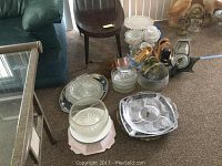 Wide view of multiple serving platters and bowls placed on floor near chair and couch. Includes a variety of metal, glass, and ceramic items.