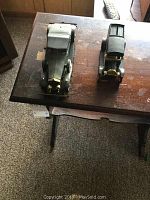 Two black vintage car coin banks on a wooden table, showing front view with details like gold accents and white wheels on Rolls Royce and side view of Ford van with text on the side.