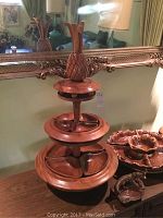 Three-tier wooden serving tray with swivel feature, pineapple handle on top, displayed on wooden furniture in room.