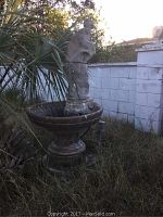 Full view of the electric water fountain with statue and basin, situated outdoors with surrounding plants and grass.