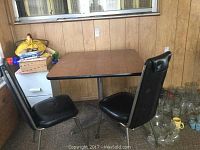 Full view of square table with two black vinyl chairs in a room with wood-paneled walls and carpeted floor. Glassware is seen beside the table.