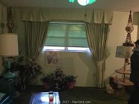 Curtains hung over window with green roller blind, showing full valance, tassels, and tiebacks. Room setting includes lamp and small shelf.