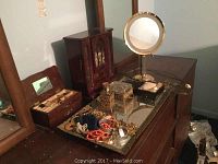 Wooden vanity mirror on dressing table with costume jewelry displayed on a mirrored tray alongside two wooden jewelry boxes and decorative perfume bottles.
