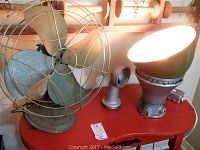 Photo showing both items on a red surface including the vintage fan and the lit Appleton vented lamp with gooseneck.