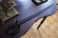 Full view of curved metal saber sword on a wooden table with an ornate metal item in background. Shows whole blade and iron guard, handle absent.