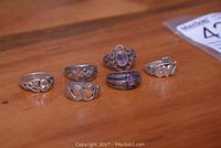 Six sterling silver rings laid out on a wooden surface showing various designs including a purple stone, Claddagh ring, and different patterns.