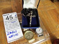 Two men's watches; one gold-tone Seiko and one Wenger, displayed with a black bead rosary on a blue cushion box and a MaxSold auction tag on wooden floor.