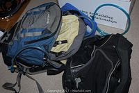 Blue and gray Camelbak hydration backpack with attached drinking tube lying on the floor with other backpacks.