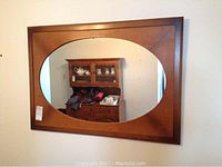 Front view of the wall mirror showing the oval glass and wooden frame mounted on a wall, reflecting furniture and room items.