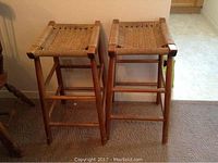 Two wooden bar stools with woven rush seats shown frontally on carpeted floor.