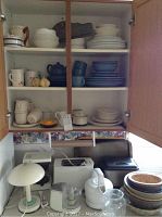 Overview of kitchen items including stacked dishes, mugs, small appliances, and lamp arranged on a counter and inside a cabinet.