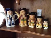 Front view of the full set showing the white and blue Toby teapot on left, brown Toby teapot second, and three Toby jugs decreasing in size from left to right.