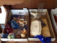 View of various collectibles laid inside two cardboard boxes including blown glass items, pottery mugs, china flowers, and other small items.