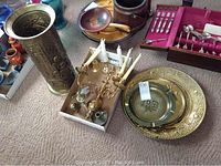 A group of brass decorative items including a tall umbrella or dress stand, candle wall sconces with candles, four brass plates, small brass figurines, and other miscellaneous brass items placed on carpet.