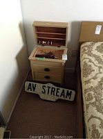 Overall view of beige sewing cabinet with hinged top open, revealing wooden spools inside, next to the AV Stream decorative street sign placed on the floor against wall.