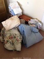 Pile of assorted bed linens including floral comforter, blue comforter, knitted throw, and multiple folded bed sheets against a wall.