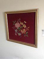 Large floral needlepoint with multicolor flowers on burgundy background in gold frame hanging on a wall, showing detail and framing