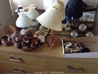 Dresser top holding 3 different table lamps, collection of 5 small teddy bears, woven basket, framed cross stitch, vintage landline phone, and wooden barometer with clock and thermometer.