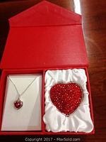 Red box containing heart-shaped sterling silver necklace with pink synthetic stone pendant and heart-shaped compact adorned with ruby colored stones