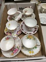 Six assorted china cup and saucer sets arranged in a box showing various floral patterns and styles.