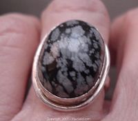 Front angle close-up of oval snowflake obsidian cabochon ring worn on finger showing black and gray-white stone with silver band.