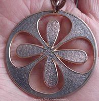 Close-up of round metal pendant showing flower petal design with light lavender enamel and textured silver-tone metal sections.
