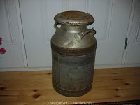 Front angle view of the vintage galvanized milk can showing the lid and one handle, on a wooden floor against a white door.