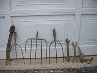 Photo of three old pitchforks with metal tines and worn handles, two large rusty wrenches, plow point, harness part, and hook all with rusty patina leaning against a white door.