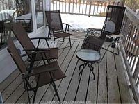 View of three brown mesh folding chairs, one gravity lounge chair, small round glass-top table and white folding resin table on wooden deck.