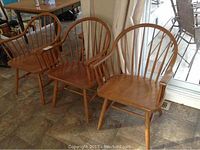 Set of three Windsor-style wooden arm chairs arranged in a row on tiled floor near window and curtains.