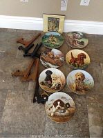 All seven collector plates laid on floor, showing various breeds of puppies including spaniel, labrador, dachshund, retriever in outdoor settings. Multiple wooden and black plastic stands beside plates. Small decorative tile also included.