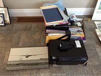 Overview photo showing HP Photosmart D110 printer, stack of assorted folders and files, framed print of a glider airplane on board, and assorted papers on tiled floor.