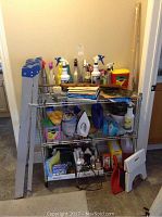 Full view showing the aluminum step ladder on the left, metal wire shelving with multiple cleaning products, iron, cleaning tools, dustpan, folding stool on the floor.