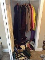 Image showing several medium-sized ladies coats and jackets in multiple colors on hangers inside a closet, with footwear and miscellaneous items on the floor below.