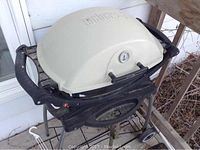 Beige Weber grill with lid closed on wheeled cart against house siding