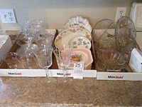 Various stemware glasses, decorative plates including Johnson Bros dessert plates, and other clear glassware arranged in boxes on countertop.