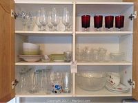 Upper kitchen cupboard displaying various glassware and bowls. Top left has clear stemware with blue floral designs. Top right red glasses. Middle left stacked bowls and plates. Middle right small clear bowls. Bottom left clear glasses including some with color. Bottom right large clear glass salad bowl and small plates.