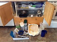 Photo showing open wooden cabinet with various kitchen items inside and on the floor in front, including stainless steel mixing bowls, plastic containers, black baking tray, dish rack with plates, and small metal bowls.