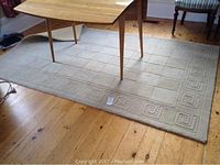 Photo showing full view of beige wool area rug under a wooden table on a wood floor. Rug has square grid central pattern and Greek key border pattern.