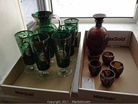 Amber glass decanter with silver overlay and 5 matching glasses next to green glass lemonade jug and 6 matching glasses, all displayed in cardboard boxes.