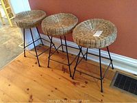 Three stools with black metal frames and round woven wicker seats placed side by side on a wood floor against a red wall.