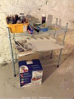 Full view of metal wire shelf with yellow container of paintbrushes, miscellaneous paint supplies, craft boards and paper sheets on middle shelf, and Nesco dehydrator box on floor beneath.