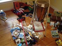 Overall view of lot showing metal garbage pail with tripod stand, assorted cleaning supplies, and folded fabrics on wood floor.