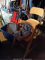 Photo showing wooden folding high chair with cushion, blue plastic tote bin, red rolling grocery cart, and other items in background.