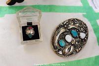 Photo showing the silver ring in a clear plastic display case and the silver belt buckle with turquoise and white stones on a white background with green tape.
