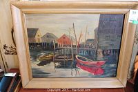 Framed oil painting showing a dock with boats in the water and wooden buildings in the background, signed lower left corner.