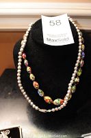 Two necklaces displayed on a black stand: a sterling silver bead necklace and a colorful Millefiori glass bead necklace.