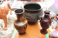 Photo showing three items on a wooden surface: two bronze vases and one enamelled planter with colorful decoration.