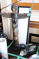 Wood totem pole with spread wings painted in black, brown, red, and beige with visible chips, displayed indoor next to a framed white embossed artwork and a black and white loon decoy.